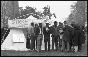 Strikers and supporters milling about outside a tent, a woman and young men look over a headline in the newspaper, statue of Lafayette in the background