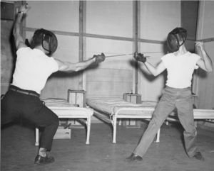 Two Suffolk University students fence on stage with cots in background, possibly a theatre performance