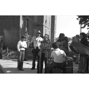 Older woman speaks into microphone at a Latino street festival, with onlookers listening and musicians waiting to play music