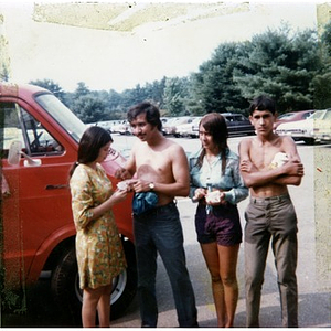 Two male and two female Latino youths stand next to a red van in the parking lot adjacent to an unidentified park and lake, while on a summer day trip sponsored by La Alianza Hispana.