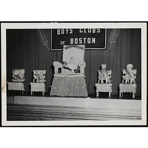 Five dolls and sashes placed in chairs on the stage for a Boys' Club Little Sister Contest award ceremony