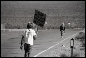 Activist on the road near the entrance to the Nevada Test Site holding a sign reading 'Stop nuclear weapons testing', wearing a t-shirt reading 'Nukes Het!': Nevada Test Site peace encampment