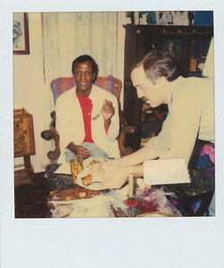 A Photograph of Marsha P. Johnson Sitting with a Fork In Her Hand, Eating Food a Coffee Table