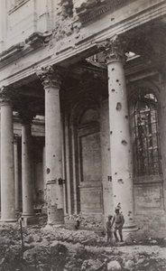 Two soldiers standing in front of a damaged building with columns pockmarked from artillery fire , Arras