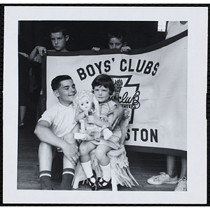 The Little Sister Contest winner holds a doll while posing with her brother in front of the Boys' Clubs of Boston banner