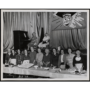 A row of women pose with a display of baked goods as two women look on from a stage in the background during a Mothers' Club event