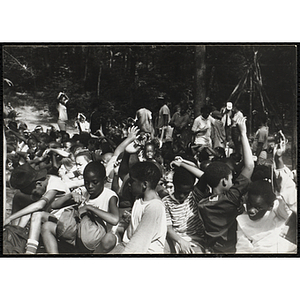 A group of children sitting in the woods raise their hands