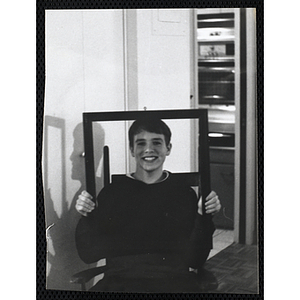 A teenage boy holding a blank frame in front of his face while sitting in a chair