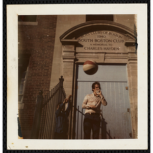 A boy throwing a basketball into the air while standing with a man in front of the entrance to the South Boston Boys' Club