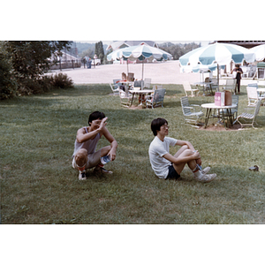 Two young men sit in the grass near a cluster of tables with green and white umbrellas