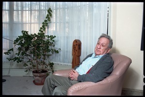 Fine art photographer Aaron Siskind: portrait with a potted plant in the background, while seated in a armchair in his living room