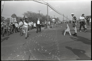 Antiwar demonstration at Fort Dix, N.J.: line of protesters marching past barbed wire and military police barricade (one raising fist in Black Power salute)