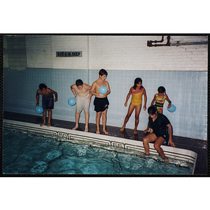 Children with balloons stand and sit poolside in a natorium