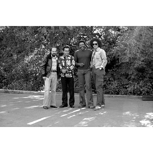 Full-length portrait of four Hispanic men, standing in front of trees and shrubs, facing front, at a Latino street festival