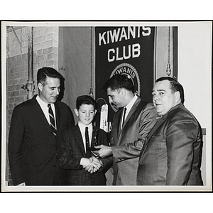 A man presents a trophy to a boy and shakes his hand while two other men stand beside them at a Charlestown Kiwanis Club awards event