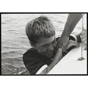 A boy hangs off the side of a sailboat as a man holds him by the arm in Boston Harbor