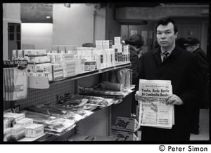 Bill Baird, contraception rights advocate, standing in a pharmacy and holding up the day's newspaper next to pharmacy supplies