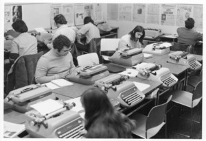 Overhead shot of Suffolk University students working at typewriters, newspapers hanging on walls