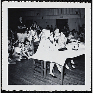 The panel of judges sit at a table while the audience sits and stands behind them during a Boys' Club Little Sister Contest