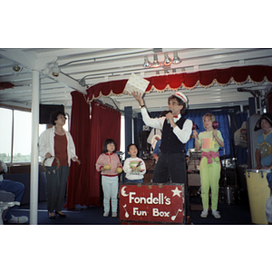 Children stand on a stage while a man speaks into a microphone, behind a sign that reads, "Fondell's Fun Box"