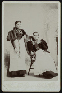 Two unidentified women, one seated and one standing, full length, facing front, location unknown, undated