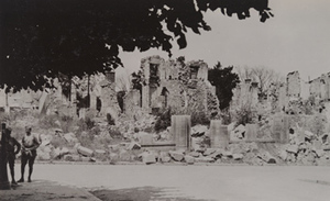 Soldiers standing in the shade of a tree, with destroyed town buildings in the background