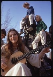 Judy Collins seated at the base of a statue, playing guitar for children