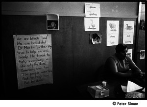 Teacher seated at a table at the Liberation School: in front of a poster honoring Martin Luther King: 'He tried to help everbody in the city. He died. Somebody shot him...'