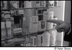 Bill Baird, contraception rights advocate, standing in a pharmacy, pointing at contraceptives in a medicine case and holding up the day's newspaper (close-up of Baird's hand)
