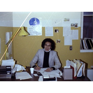 Male employee with Afro haircut, seated behind a desk, writing, at La Alianza Hispana offices, Roxbury, Mass.