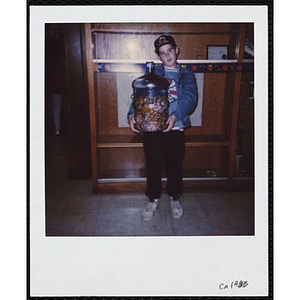 A boy holding a water cooler bottle filled with candy and posing in front of a display case at the Boys & Girls Club