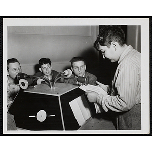 A boy reads into a microphone on a console in a radio station as three other boys look on
