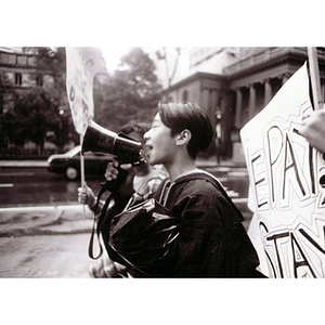 Young man speaks into a bullhorn while two people hold up protest signs at an unidentified demonstration