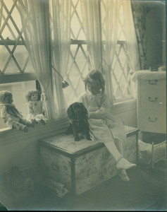 Portrait of a young girl, seated on a chest, facing left, holding a dog, location unknown, undated