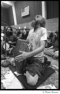 Ram Dass seated on a floor with a drum during an appearance at the College of Marin, chanting