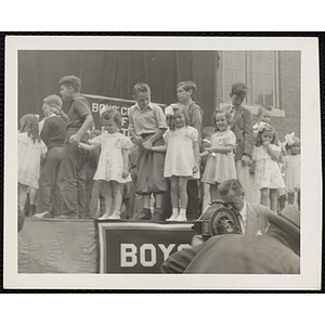Boys hold hands with their sisters on the outdoor stage while a cameraman, facing left, operates a motion picture camera during a Boys' Club Little Sister Contest