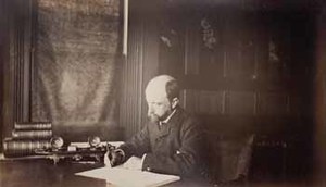 Henry Adams seated at desk in dark coat, writing