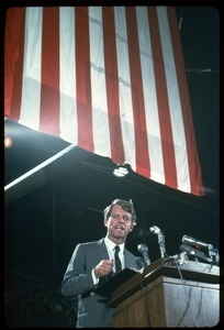 Robert F. Kennedy on the campaign trail, speaking beneath a giant U.S. flag while stuming for Democratic candidates in the northern Midwest