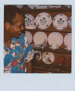 A Photograph of Marsha P. Johnson in a Blue Hawaiian Shirt Looking at Porcelain Plates