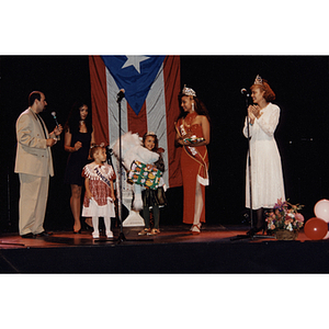 A man with a microphone speaks on stage next to three women and two little girls at the Festival Puertorriqueño
