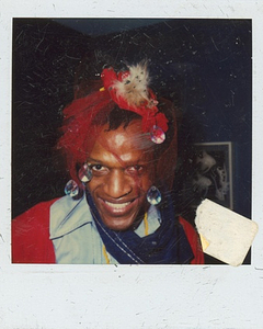 A Photograph of Marsha P. Johnson Smiling at the Camera Wearing a Red Mesh Headpiece with Large Gems