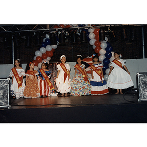 Seven little girls stand on stage in dresses and sashes at the Festival Puertorriqueño