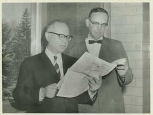 John Sharp Harris standing indoors, reading paper, with man