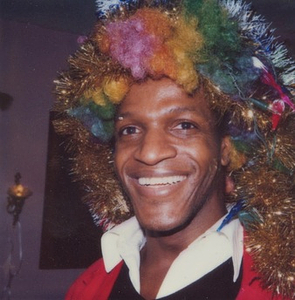 A Photograph of Marsha P. Johnson Wearing a Rainbow Wig and Smiling at the Camera