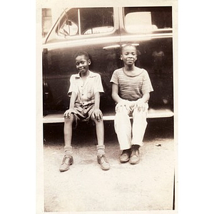 Two unidentified boys sit against a black vehicle