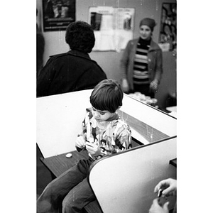 Hispanic American boy sits on top of a desk in a classroom at La Alianza Hispana, Roxbury, Mass