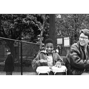 Little boy playing bongo drums alongside Alex Alvear as they sit outside on a park bench.