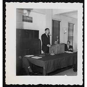 A man standing behind a podium while two others look on during the Board of Overseers meeting