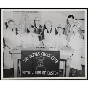 Members of the Tom Pappas Chefs' Club pose with their club banner and three unidentified adults in a kitchen
