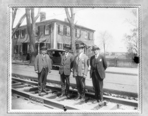 Four men in suits standing on tracks in front of a house and car
